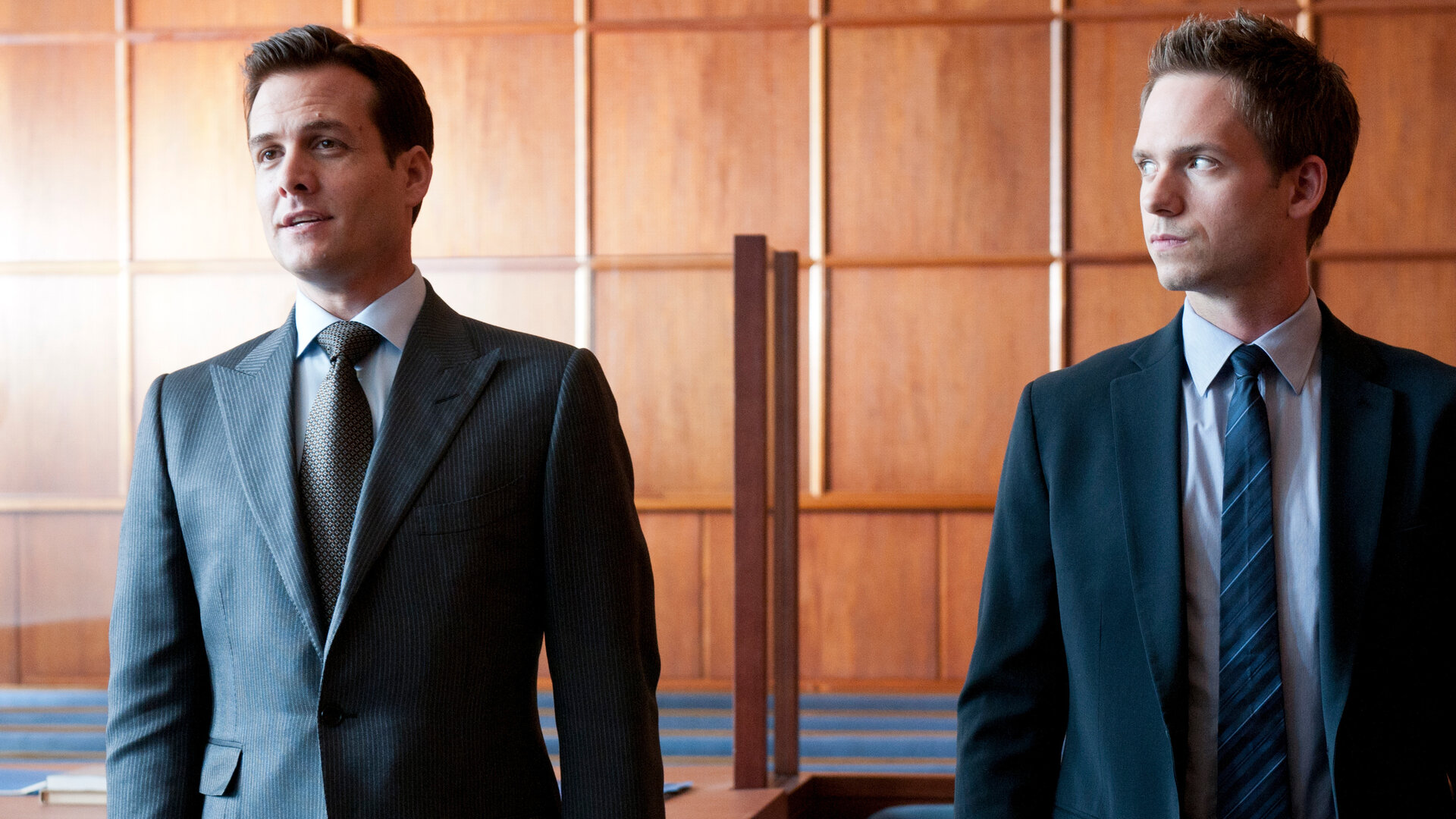 "Suits - Enthüllungen": Harvey (Gabriel Macht) und Mike (Patrick J. Adams) stehen nebeneinander im Gerichtssaal. Beide tragen Anzüge und blicken in unterschiedliche Richtungen. Im Hintergrund ist eine hölzerne Wandverkleidung zu sehen.