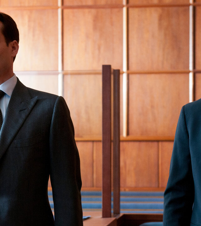 "Suits - Enthüllungen": Harvey (Gabriel Macht) und Mike (Patrick J. Adams) stehen nebeneinander im Gerichtssaal. Beide tragen Anzüge und blicken in unterschiedliche Richtungen. Im Hintergrund ist eine hölzerne Wandverkleidung zu sehen.