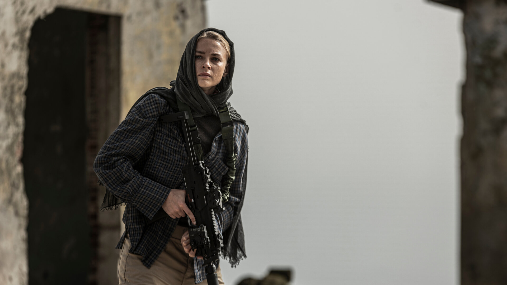 "Kabul - Flucht aus der Hölle: Ein riskanter Plan": Vera (Jeanne Goursaud) tritt mit einem Sturmgewehr bewaffnet aus den Ruinen eines zerfallenen Gebäudes. Sie trägt ein Kopftuch und eine karierte Jacke, ihr Blick ist konzentriert und angespannt, während sie die Umgebung absichert.