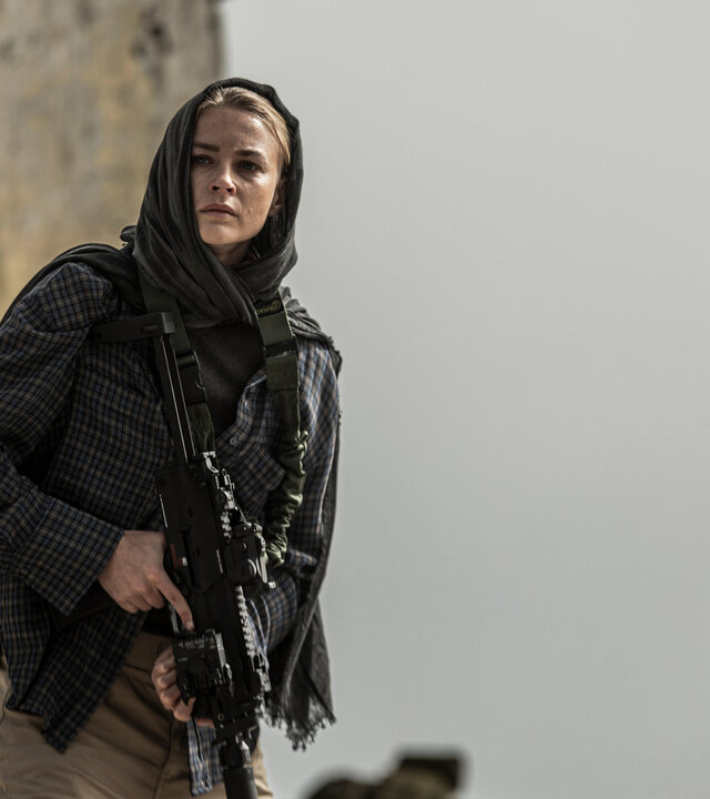 "Kabul - Flucht aus der Hölle: Ein riskanter Plan": Vera (Jeanne Goursaud) tritt mit einem Sturmgewehr bewaffnet aus den Ruinen eines zerfallenen Gebäudes. Sie trägt ein Kopftuch und eine karierte Jacke, ihr Blick ist konzentriert und angespannt, während sie die Umgebung absichert.