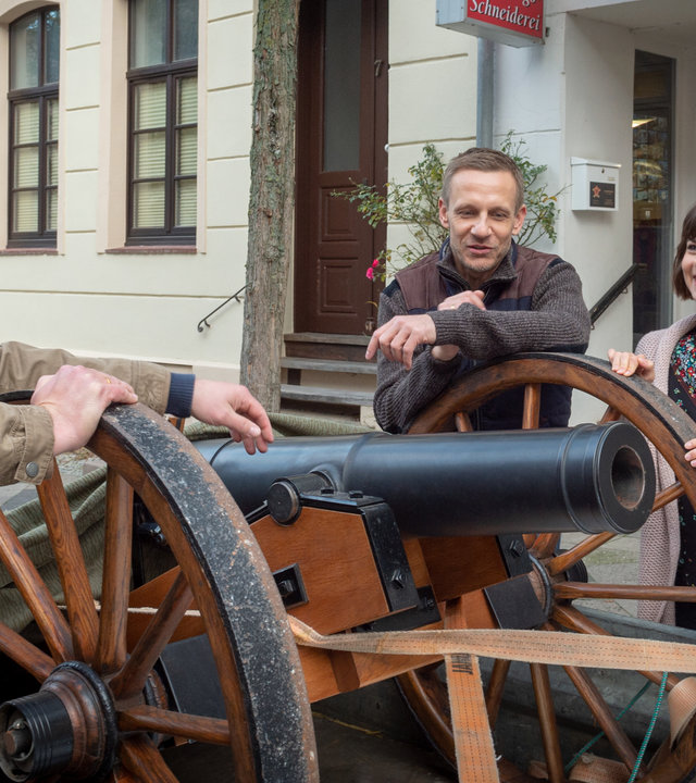 "SOKO Wismar – Tödlicher Salut": Vor einer Schneiderei steht eine historische Kanone auf einem Anhänger. Achim Buchholz (René Schwittay) stützt sich lässig darauf ab. Hinter der Kanone stehen Oliver (Mike Hoffmann) und Lilo Michel (Sandra Julia Reils). Lilo strahlt Achim an.