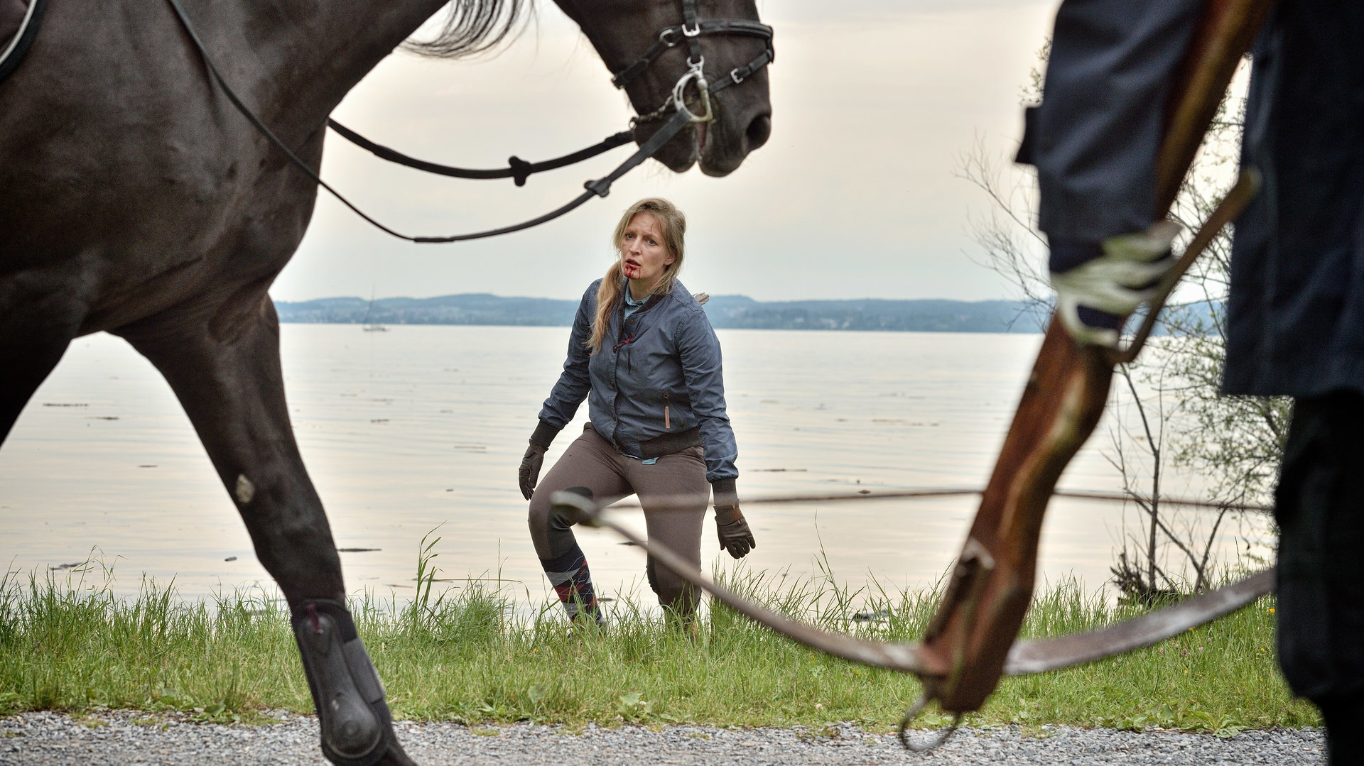 "Die Toten vom Bodensee - Blutritt": Am linken Bildrand ist ein Pferd zu erkennen. Im Anschnitt ist eine Person zu erkennen, die mit Handschuhen eine Armbrust nach unten hält. Im Hintergrund bewegt sich Marlene Stöhr (Stefanie von Poser) auf das Pferd und die Person zu. Hinter ihr befindet sich der Bodensee. Die Frau trägt Reitkleidung. es ist zu erkennen, dass etwas aus ihrem Rücken ragt. Aus ihrem Mund läuft Blut.