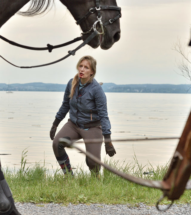 "Die Toten vom Bodensee - Blutritt": Am linken Bildrand ist ein Pferd zu erkennen. Im Anschnitt ist eine Person zu erkennen, die mit Handschuhen eine Armbrust nach unten hält. Im Hintergrund bewegt sich Marlene Stöhr (Stefanie von Poser) auf das Pferd und die Person zu. Hinter ihr befindet sich der Bodensee. Die Frau trägt Reitkleidung. es ist zu erkennen, dass etwas aus ihrem Rücken ragt. Aus ihrem Mund läuft Blut.