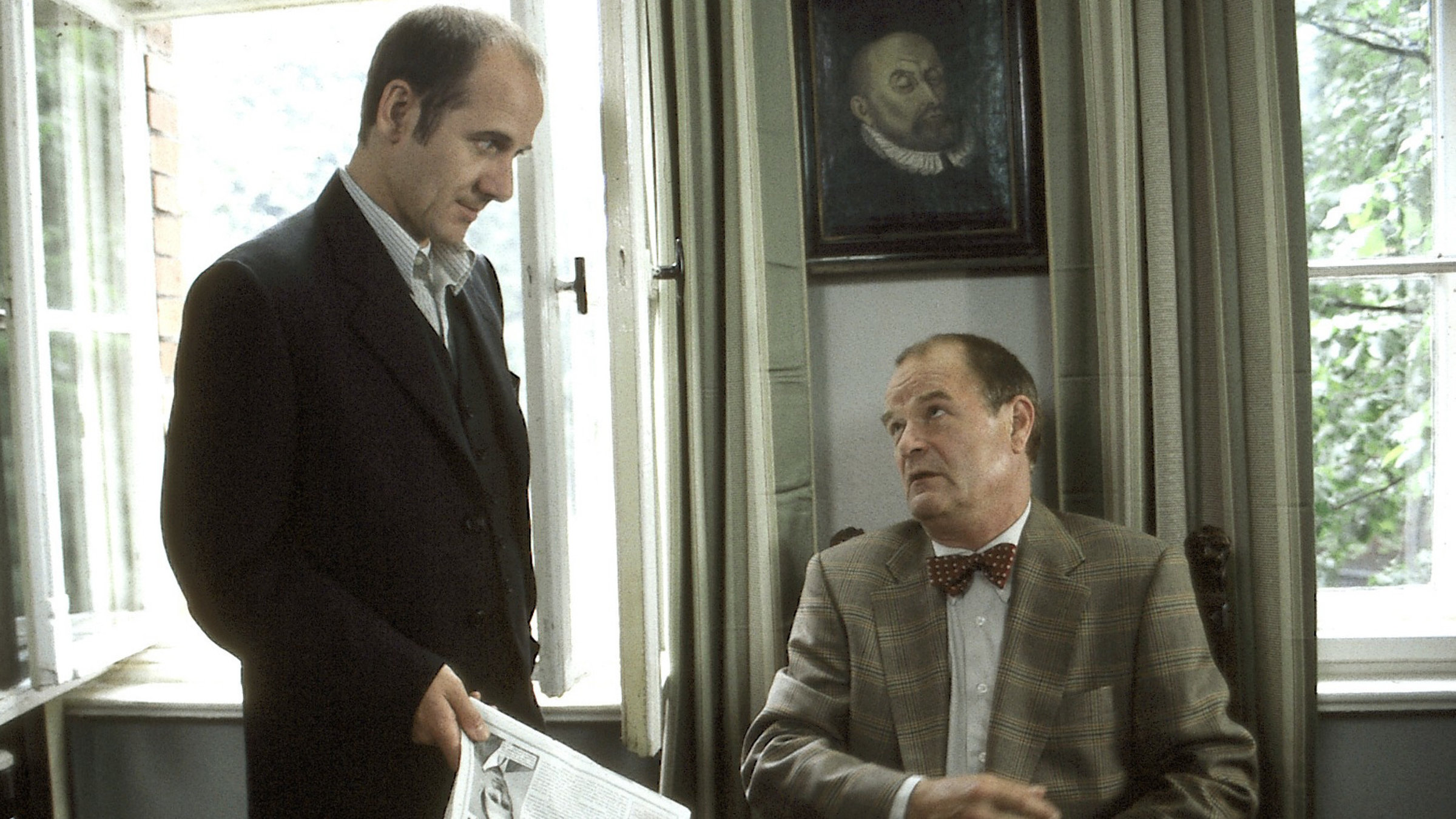 "Der letzte Zeuge - Die Fliegen, Die Maden, Der Tod": Prof. Dr. Bondzio (Dieter Mann) sitzt an seinem Schreibtisch. Dr. Robert Kolmaar (Ulrich Mühe) steht daneben und hat eine Zeitung in der Hand.