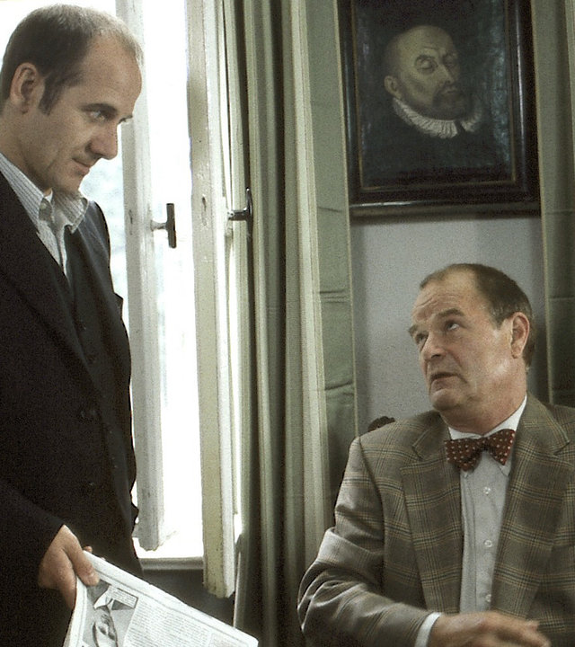 "Der letzte Zeuge - Die Fliegen, Die Maden, Der Tod": Prof. Dr. Bondzio (Dieter Mann) sitzt an seinem Schreibtisch. Dr. Robert Kolmaar (Ulrich Mühe) steht daneben und hat eine Zeitung in der Hand.