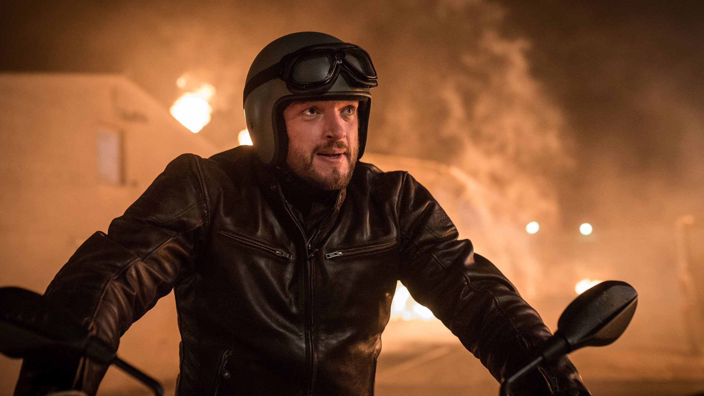 "Dengler - Am zwölften Tag": Georg Dengler (Ronald Zehrfeld) sitzt in Motorradkluft auf seinem Motorrad, im Hintergrund sieht man eine brennende Scheune.