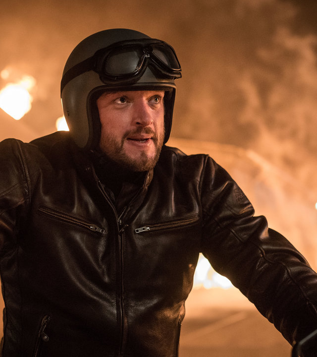 "Dengler - Am zwölften Tag": Georg Dengler (Ronald Zehrfeld) sitzt in Motorradkluft auf seinem Motorrad, im Hintergrund sieht man eine brennende Scheune.