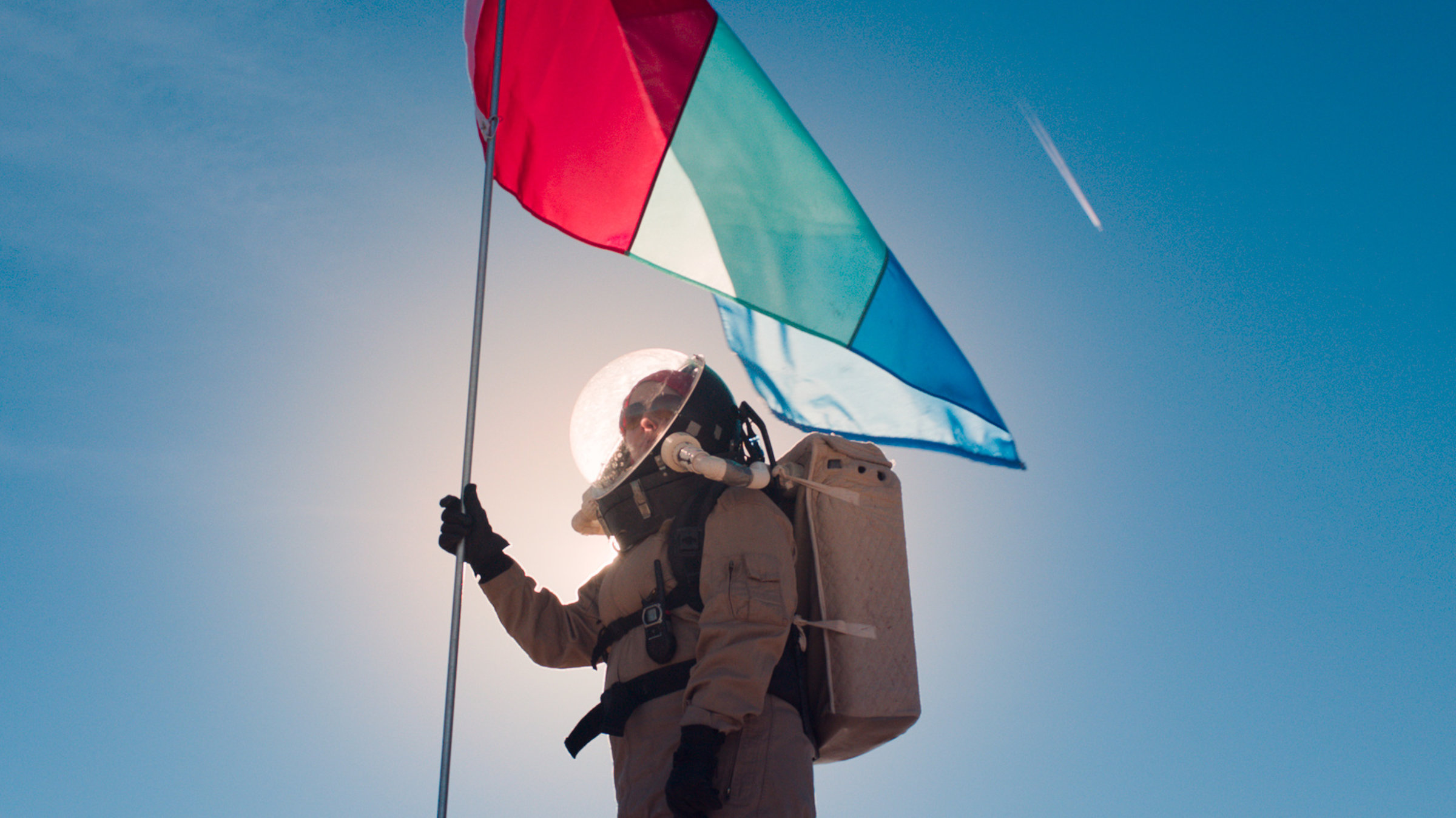 "Above and Below": Frau im Raumanzug steht im grellen Sonnenlicht auf einem Hügel und hält an einer langen Stange eine Flagge in der Hand.
