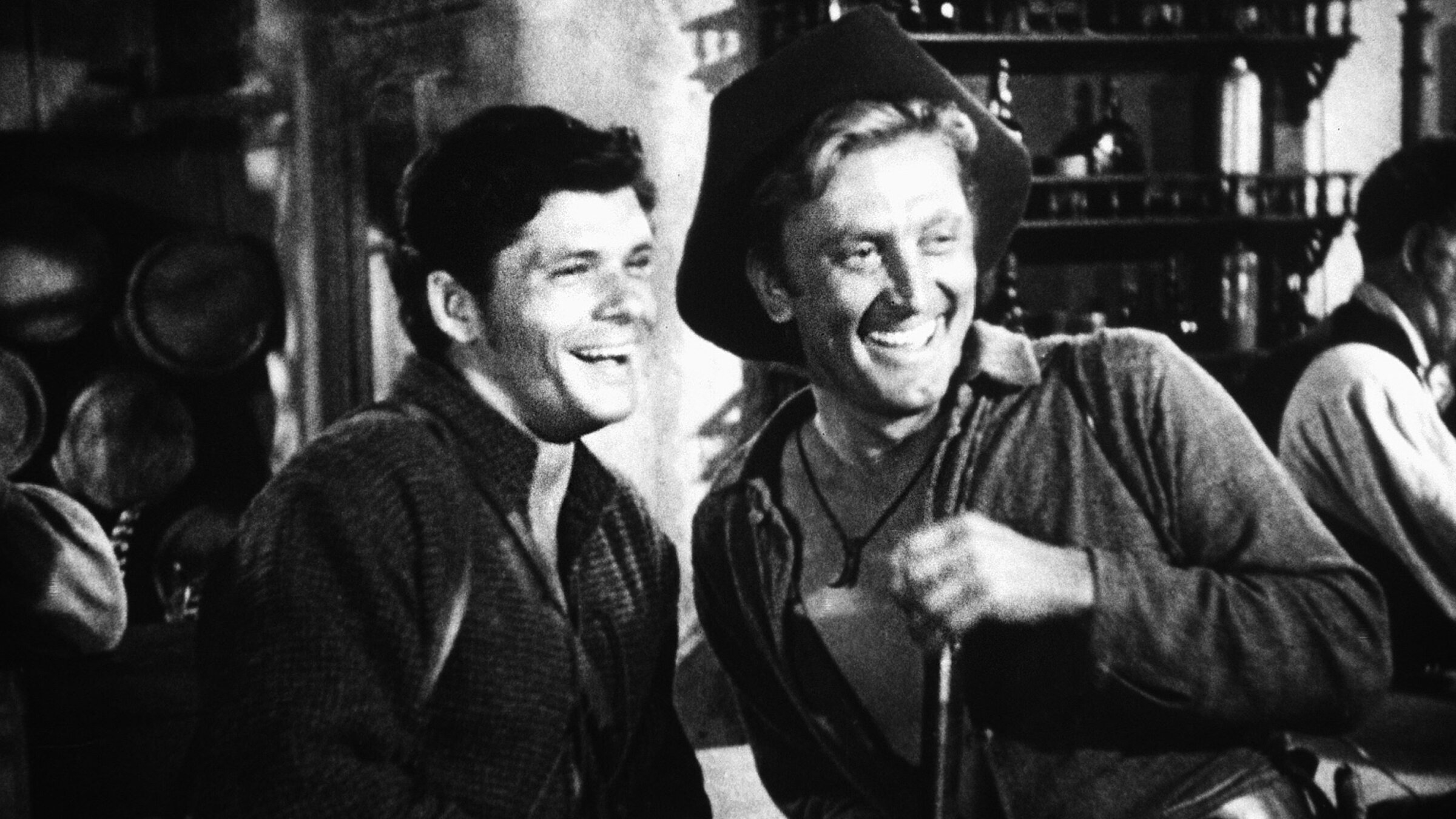 "Der weite Himmel": Jim Deakins (Kirk Douglas, rechts) und Boone Caudill (Dewey Martin) stehen in Cowboy-Kluft in einem Saloon lachend nebeneinander.