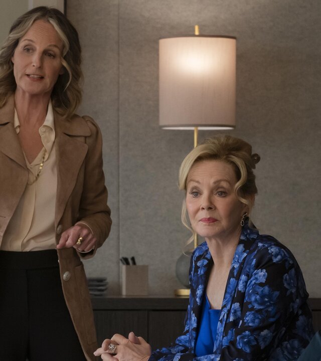 "Hacks - Bombensicher": Deborah (Jean Smart) sitzt in einer blau gemusterten Bluse neben Jimmy (Paul W. Downs) in einem Raum. Neben ihr steht Winnie Landell (Helen Hunt) die gestikulierend spricht.