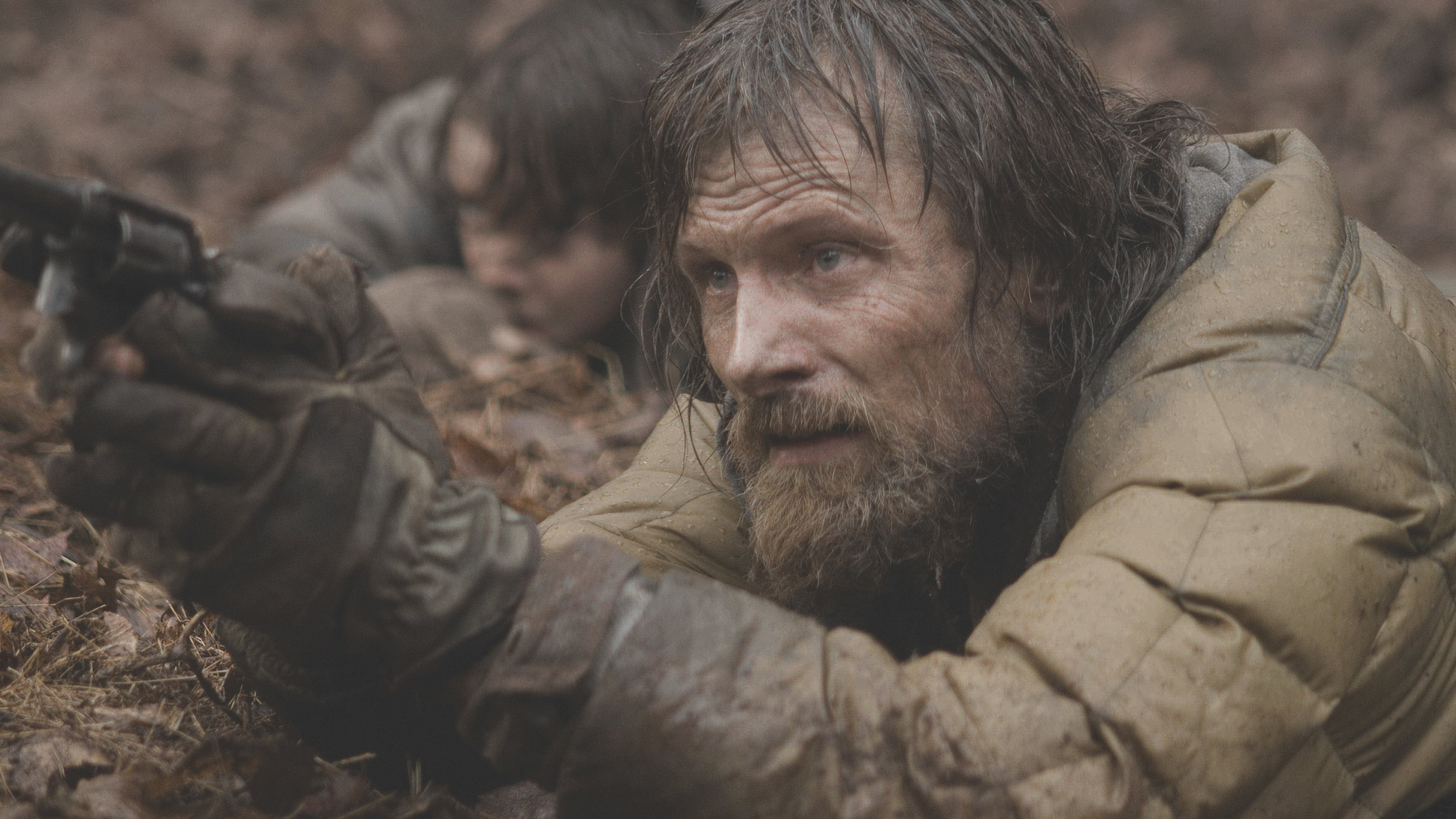 "The Road": Ein Mann (Viggo Mortensen) mit zerzaustem Bart und in einer dreckigen grün-braunen Daunenjacke kauert sich hinter einem erdigen Abhang und hält in beiden Händen eine Pistole vor sich, während er konzentriert nach vorn schaut. Hinter ihm, fast unscharf, ist der Kopf eines Jungen (Kodi Smit-McPhee) zu sehen.