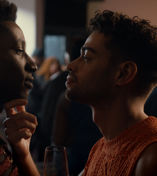 "House of Bellevue - Sex Siren": Emm (Ricco-Jarret Boateng) lehnt an der Wand und sieht João (Robin Cadet) tief in die Augen, der nah vor ihm steht und sein Kinn mit den Fingern festhält.