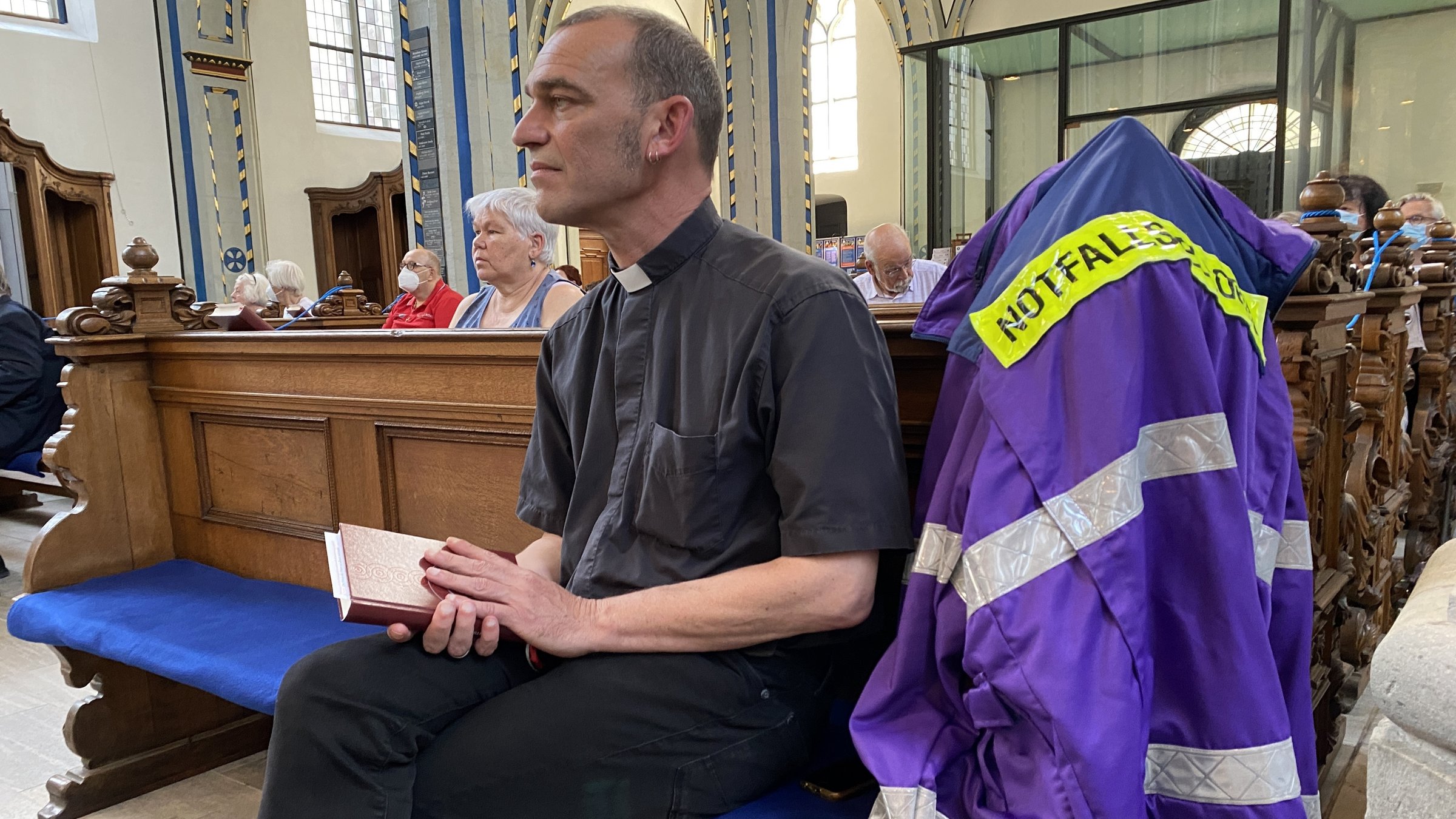 "Leben ist mehr! Plötzlich ist alles verloren - Zum Buß- und Bettag 2021": Albrecht Roebke sitzt in einer Kirche auf einer Holzbank. Er hält ein Gesangsbuch in der Hand und schaut nach rechts vorne. An der Bank hängt eine blaue Jacke mit Aufschrift "Notfallseelsorge".
