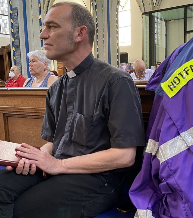 "Leben ist mehr! Plötzlich ist alles verloren - Zum Buß- und Bettag 2021": Albrecht Roebke sitzt in einer Kirche auf einer Holzbank. Er hält ein Gesangsbuch in der Hand und schaut nach rechts vorne. An der Bank hängt eine blaue Jacke mit Aufschrift "Notfallseelsorge".
