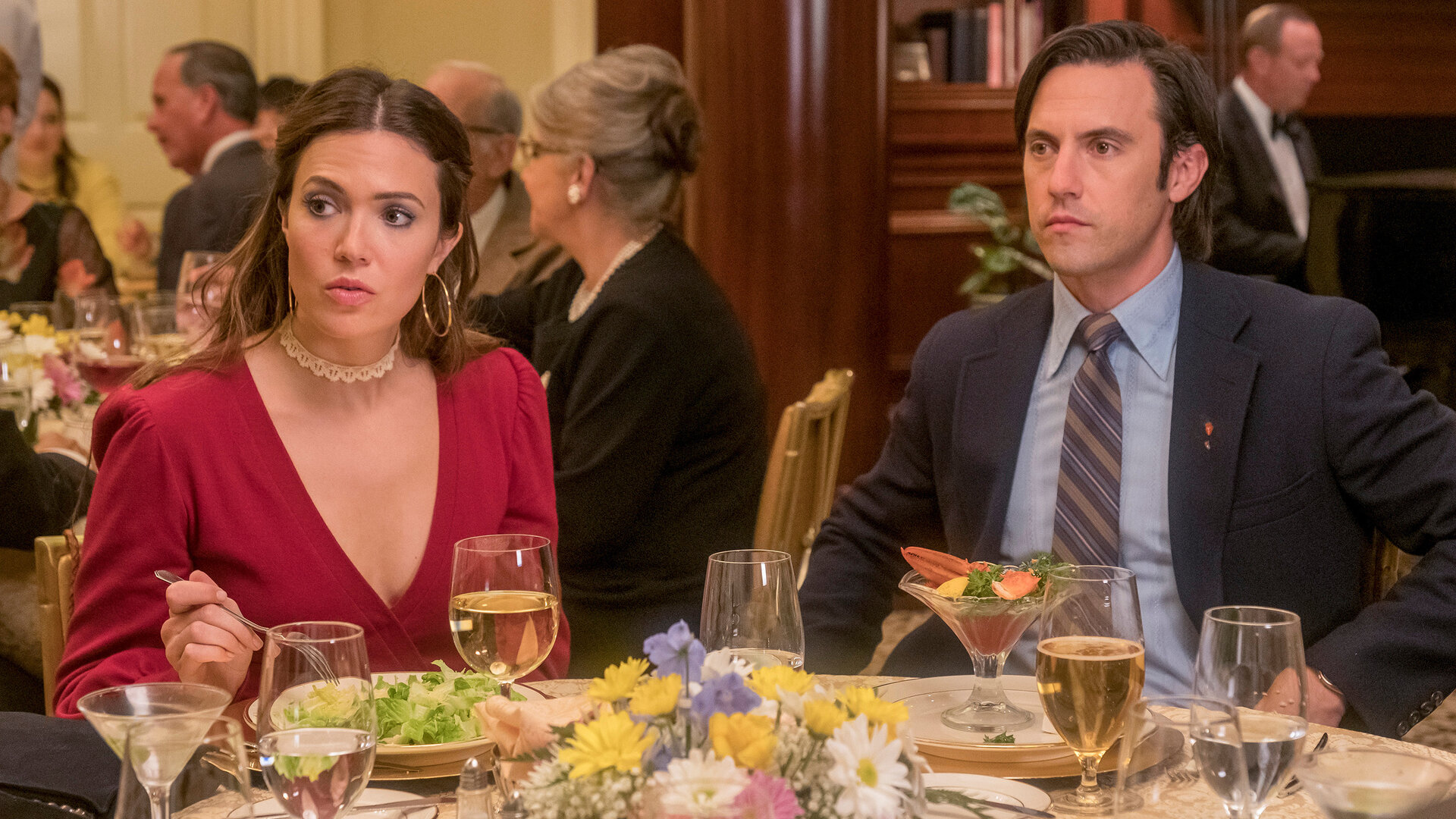 "This Is Us - Das ist Leben: Fremde": Rebecca Pearson (Mandy Moore) und Jack Pearson (Milo Ventimiglia) sitzen an einem runden Tisch in einem Restaurant. Auf dem Tisch stehen Gläser, Geschirr und verschiedene Speisen, in der Mitte befindet sich ein bunter Blumenstrauß. Beide blicken in dieselbe Richtung, doch während Rebecca fragend schaut, wirkt Jack ernst.