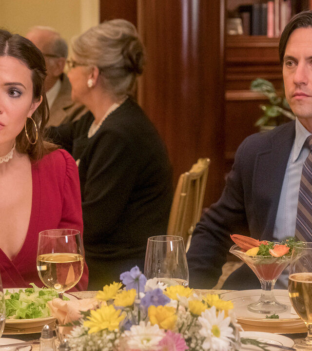 "This Is Us - Das ist Leben: Fremde": Rebecca Pearson (Mandy Moore) und Jack Pearson (Milo Ventimiglia) sitzen an einem runden Tisch in einem Restaurant. Auf dem Tisch stehen Gläser, Geschirr und verschiedene Speisen, in der Mitte befindet sich ein bunter Blumenstrauß. Beide blicken in dieselbe Richtung, doch während Rebecca fragend schaut, wirkt Jack ernst.