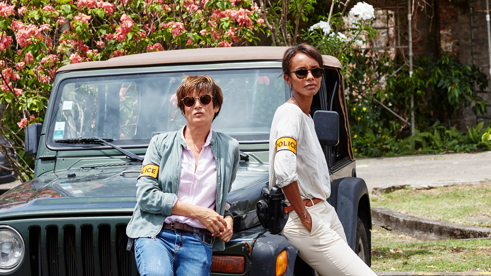 "Deadly Tropics - Willkommen auf Martinique":  Gaëlle Crivelli (Beatrice de la Boulaye) und Mélissa Sainte-Rose (Sonia Rolland) lehnen sich lässig an einen Jeep und blicken in die Ferne. Sie tragen beide eine Sonnenbrille.