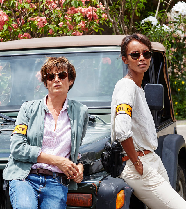 "Deadly Tropics - Willkommen auf Martinique":  Gaëlle Crivelli (Beatrice de la Boulaye) und Mélissa Sainte-Rose (Sonia Rolland) lehnen sich lässig an einen Jeep und blicken in die Ferne. Sie tragen beide eine Sonnenbrille.