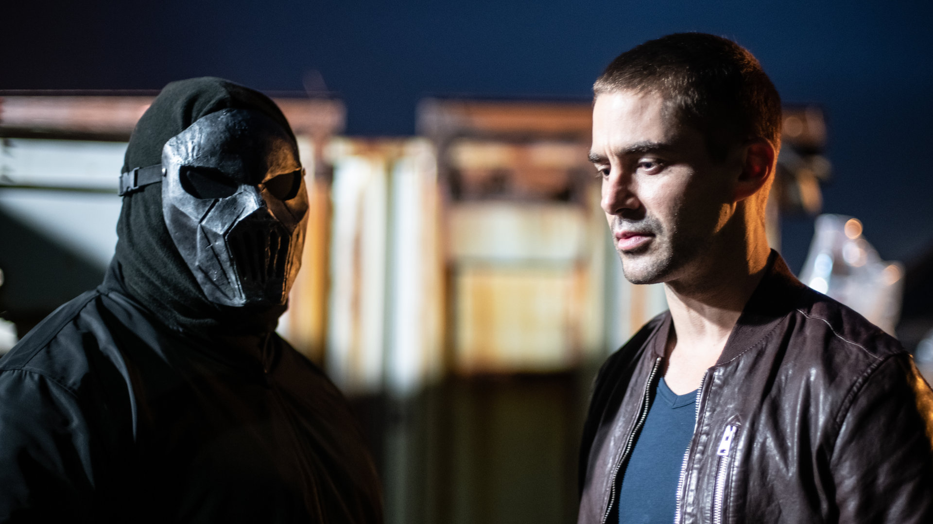 "Detective Grace - Stirb schön": Anton (Austin Hardiman) steht in seiner Lederjacke neben einem Mann, der in einen schwarzen Umhang gehüllt ist und eine schwarze Maske trägt.