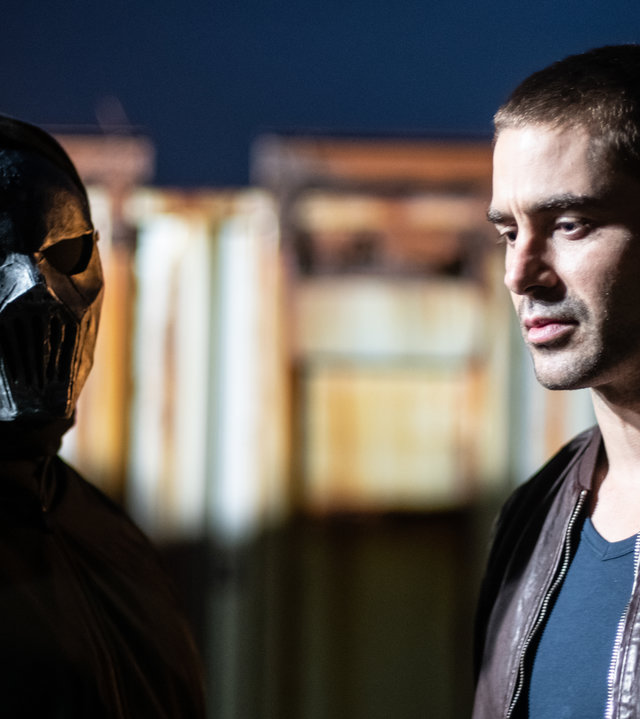 "Detective Grace - Stirb schön": Anton (Austin Hardiman) steht in seiner Lederjacke neben einem Mann, der in einen schwarzen Umhang gehüllt ist und eine schwarze Maske trägt.