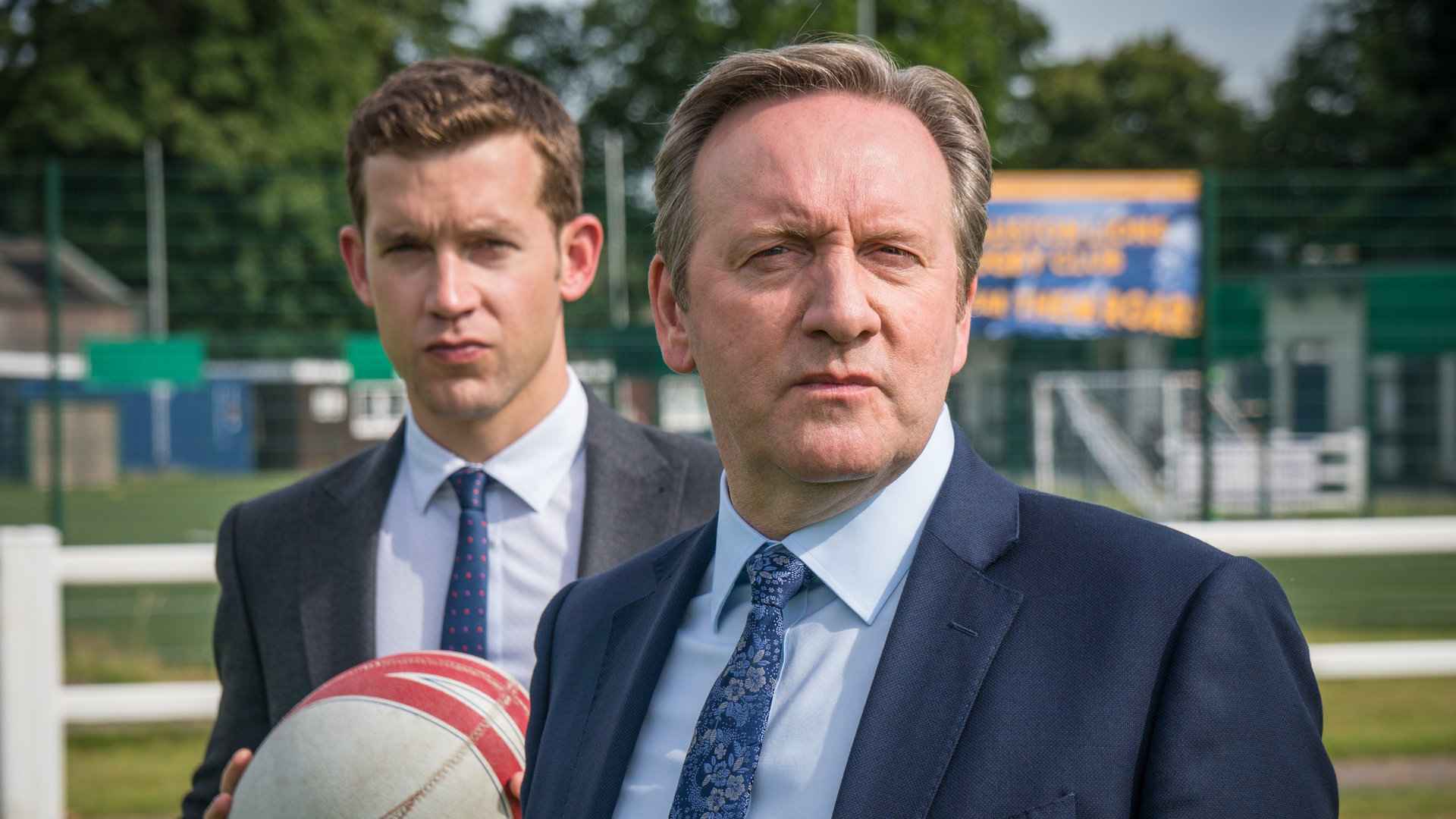 "Inspector Barnaby - Die Löwen sind los": DCI John Barnaby (Neil Dudgeon) und sein Assistent Jamie Winter (Nick Hendrix) stehen auf einem Rugby-Feld. Winter hält einen Rugby-Ball in seinen Händen.