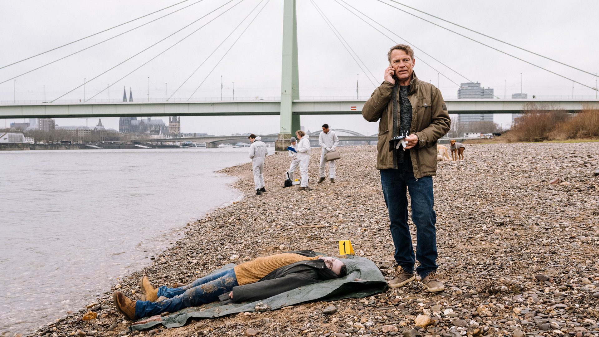 "SOKO Köln - Kinderfrei": Matti Wagner (Pierre Besson) steht neben dem toten Jan Erler (Komparse) an einem Kiesstrand und telefoniert mit ernstem Blick. Er hält einen schwarzen Gegenstand und weiße Gummihandschuhe in der rechten Hand. Der Tote liegt auf dem Rücken auf einer grauen Plane, neben ihm steht ein gelbes kleines Schild, auf dem die Zahl "eins" zu sehen ist.  Im Hintergrund stehen vier Mitarbeiter der Spurensicherung (Komparsen) und ein Hund am Strand. Hinter ihnen steht eine Brücke vor der Kulisse von Köln.