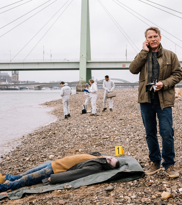"SOKO Köln - Kinderfrei": Matti Wagner (Pierre Besson) steht neben dem toten Jan Erler (Komparse) an einem Kiesstrand und telefoniert mit ernstem Blick. Er hält einen schwarzen Gegenstand und weiße Gummihandschuhe in der rechten Hand. Der Tote liegt auf dem Rücken auf einer grauen Plane, neben ihm steht ein gelbes kleines Schild, auf dem die Zahl "eins" zu sehen ist.  Im Hintergrund stehen vier Mitarbeiter der Spurensicherung (Komparsen) und ein Hund am Strand. Hinter ihnen steht eine Brücke vor der Kulisse von Köln.