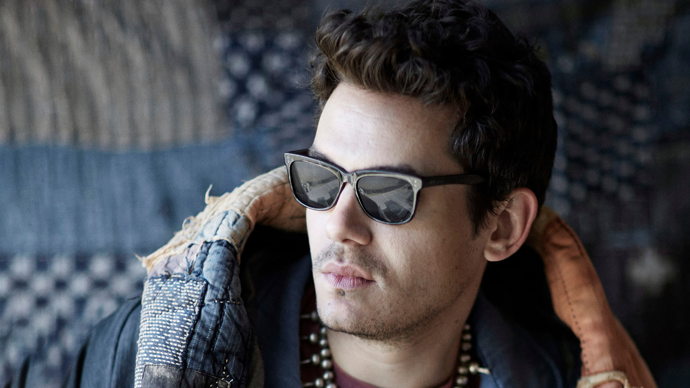 "John Mayer: Where the Light Is": Portraitfoto des Sängers und Gitarristen John Mayer. Er trägt eine schwarze Sonnenbrille und blickt seitlich an der Kamera vorbei. Über die Schultern hat er eine hellbraune Lederjacke mit Applikationen gelegt. Im Hintergrund an der Wand eine Patchwork-Decke.