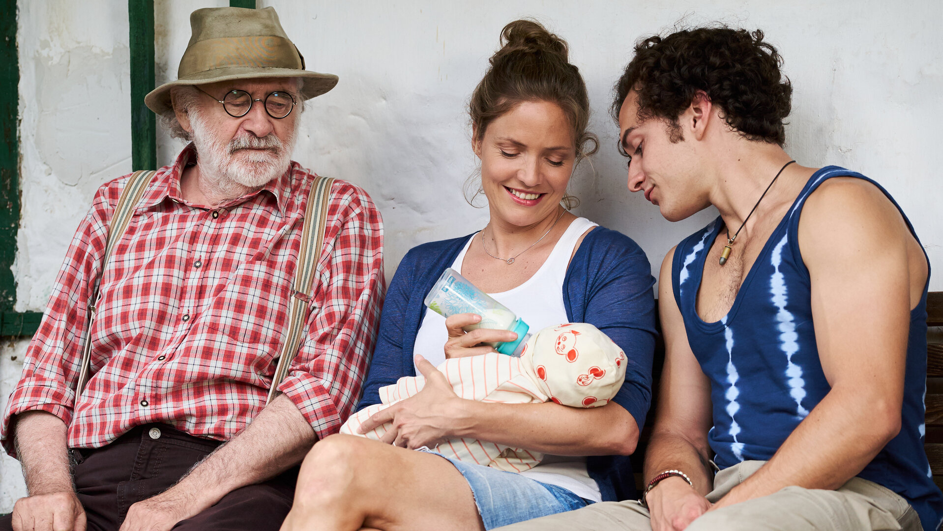 "Lena Lorenz - Mutter für drei Tage": Leo (Fred Stillkrauth) und Basti (Raban Bieling) sitzen links und rechts neben Lena (Patricia Aulitzky), die einen Säugling füttert und dabei lächelt.