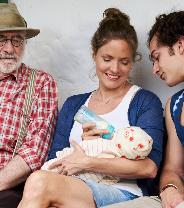 "Lena Lorenz - Mutter für drei Tage": Leo (Fred Stillkrauth) und Basti (Raban Bieling) sitzen links und rechts neben Lena (Patricia Aulitzky), die einen Säugling füttert und dabei lächelt.