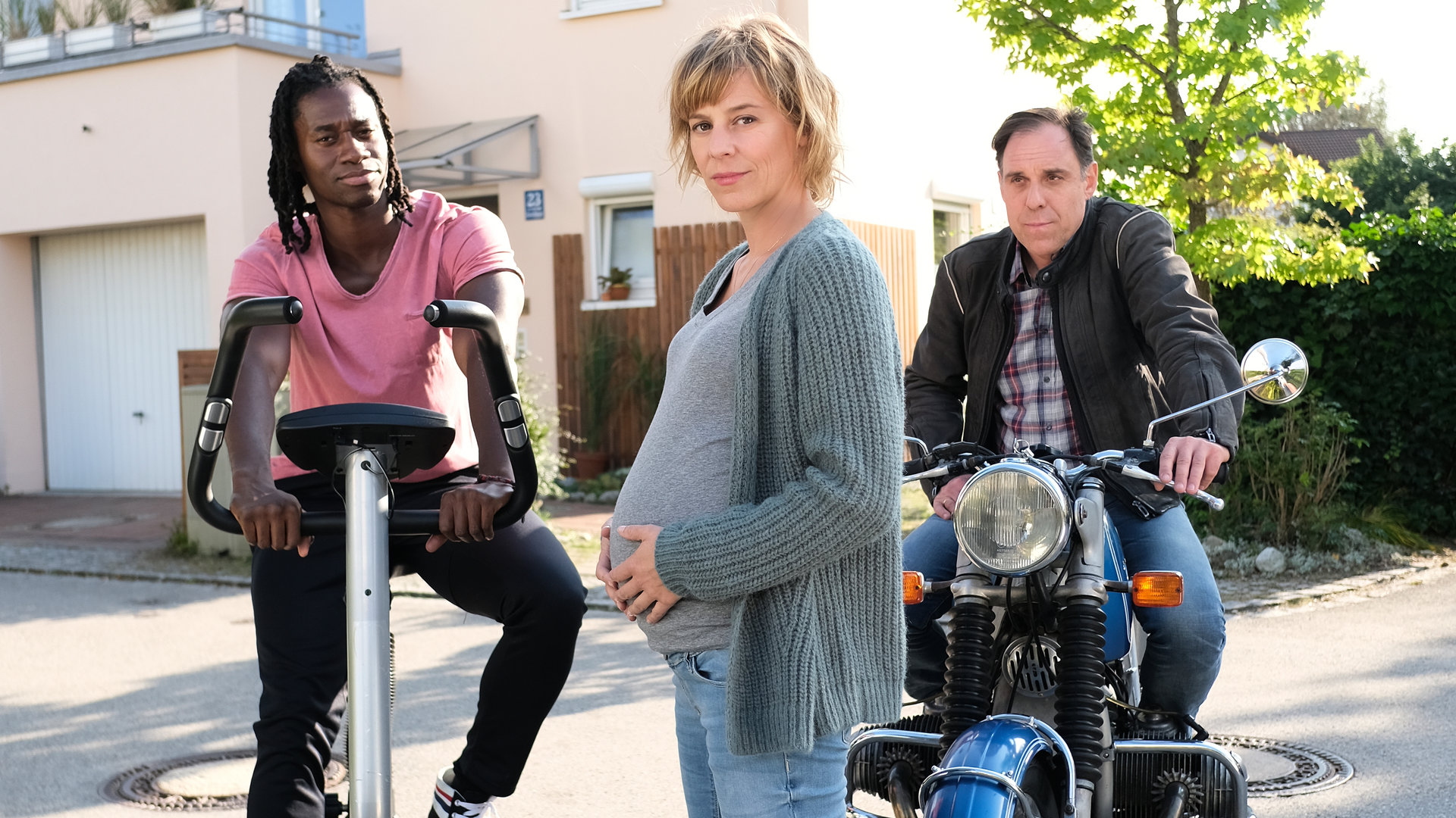 "Annie - kopfüber ins Leben": Annie (Bernadette Heerwagen) steht mit Babybauch vor ihrem Haus auf der Straße. Links neben ihr Hip-Hop-Trainer Raimund (Eugene Boateng), der auf einem Fitness-Bike sitzt. Rechts neben Annie sitzt ihr Mann Ralf (Thomas Loibl) auf einem alten Motorrad.