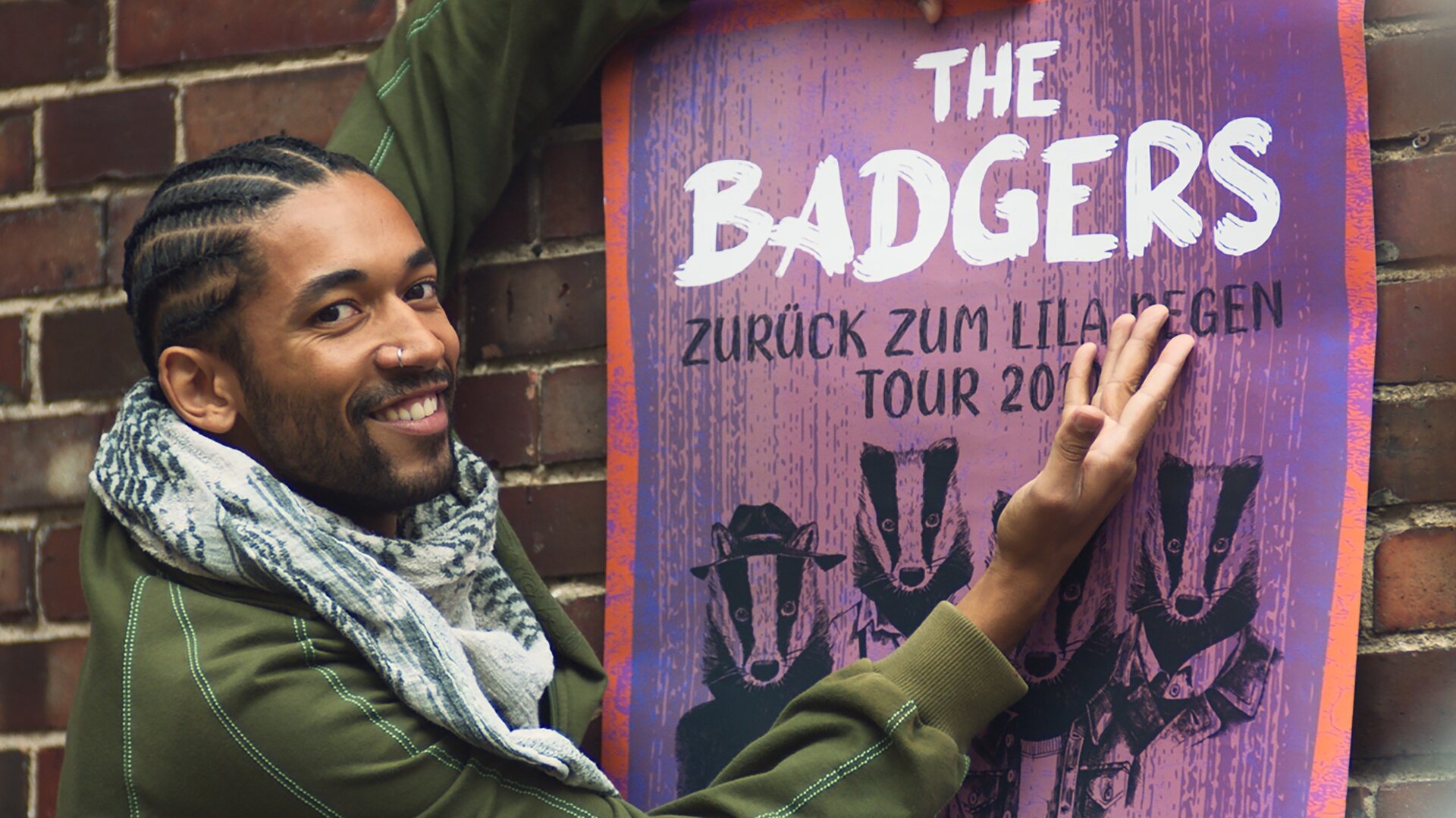 "Doppelhaushälfte - Der Traum ist aus": Theo (Benito Bause) steht stolz vor einem Bandplakat und zeigt mit seiner rechten Hand darauf. Sein Blick ist in die Kamera gerichtet.