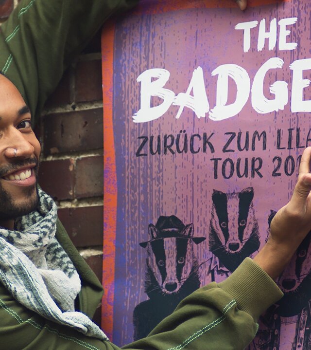 "Doppelhaushälfte - Der Traum ist aus": Theo (Benito Bause) steht stolz vor einem Bandplakat und zeigt mit seiner rechten Hand darauf. Sein Blick ist in die Kamera gerichtet.