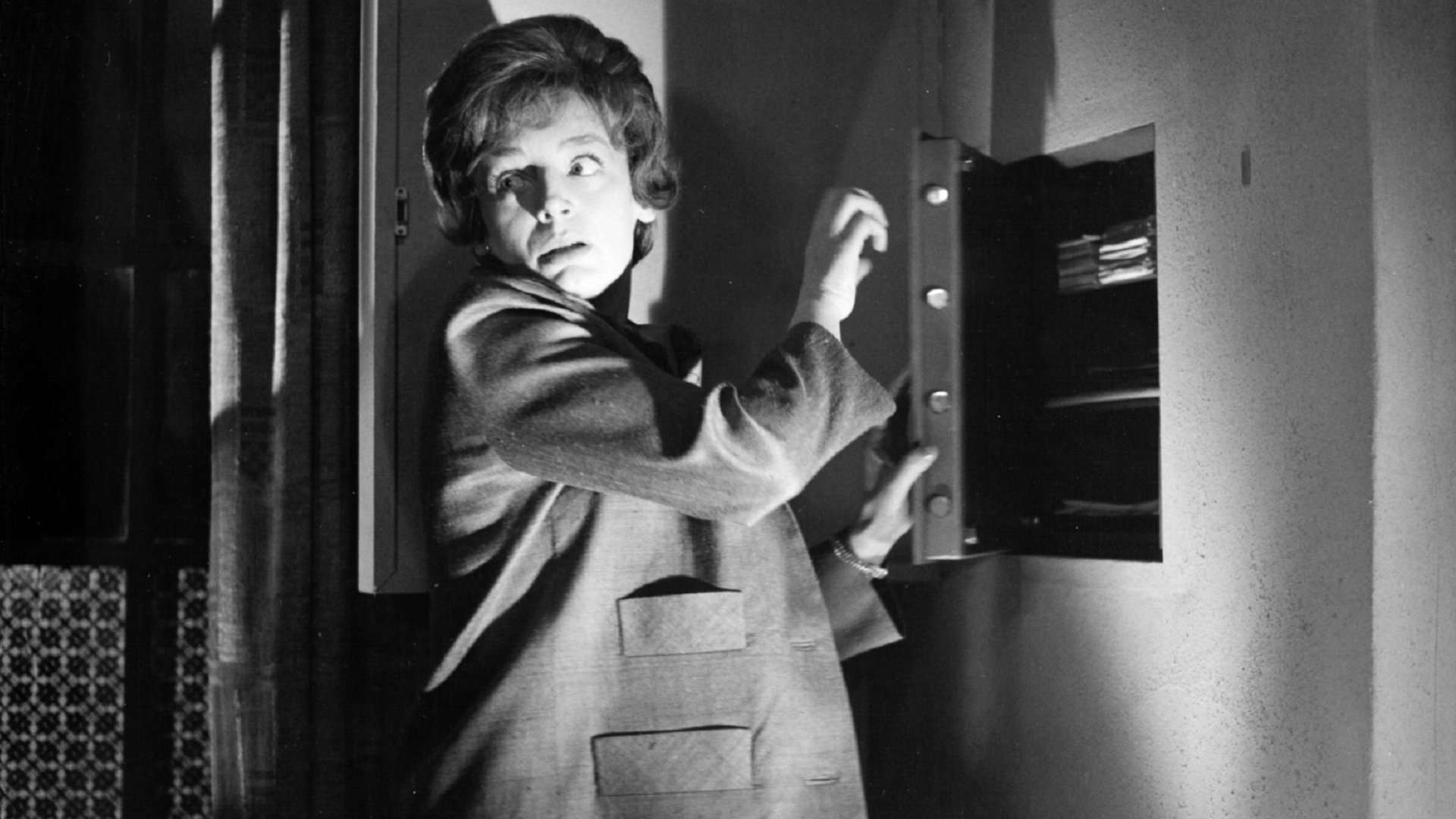 "Edgar Wallace: Der Zinker": Eine Frau steht vor einem halb geöffneten Wandsafe im Halbschatten, schlaglichtartig wird ihr Gesicht von einer einzelnen Lichtquelle erhellt. Vorsichtig blickt sie über ihre Schulter.