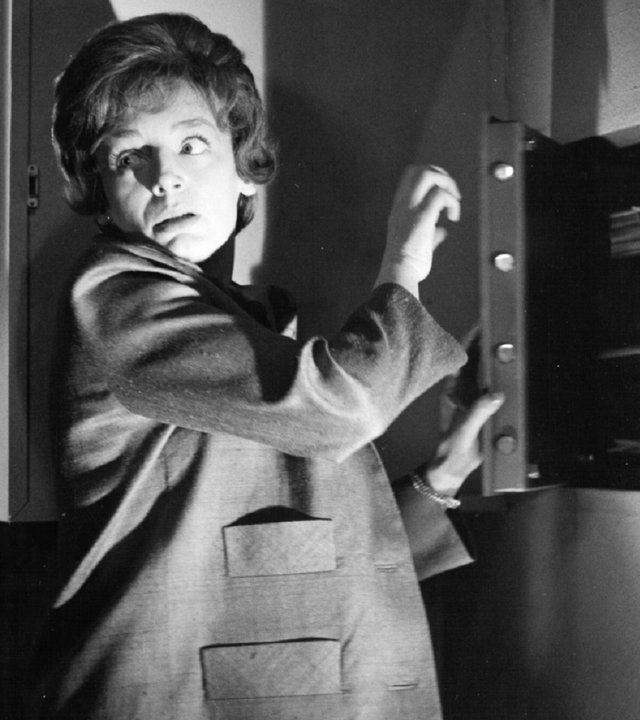 "Edgar Wallace: Der Zinker": Eine Frau steht vor einem halb geöffneten Wandsafe im Halbschatten, schlaglichtartig wird ihr Gesicht von einer einzelnen Lichtquelle erhellt. Vorsichtig blickt sie über ihre Schulter.