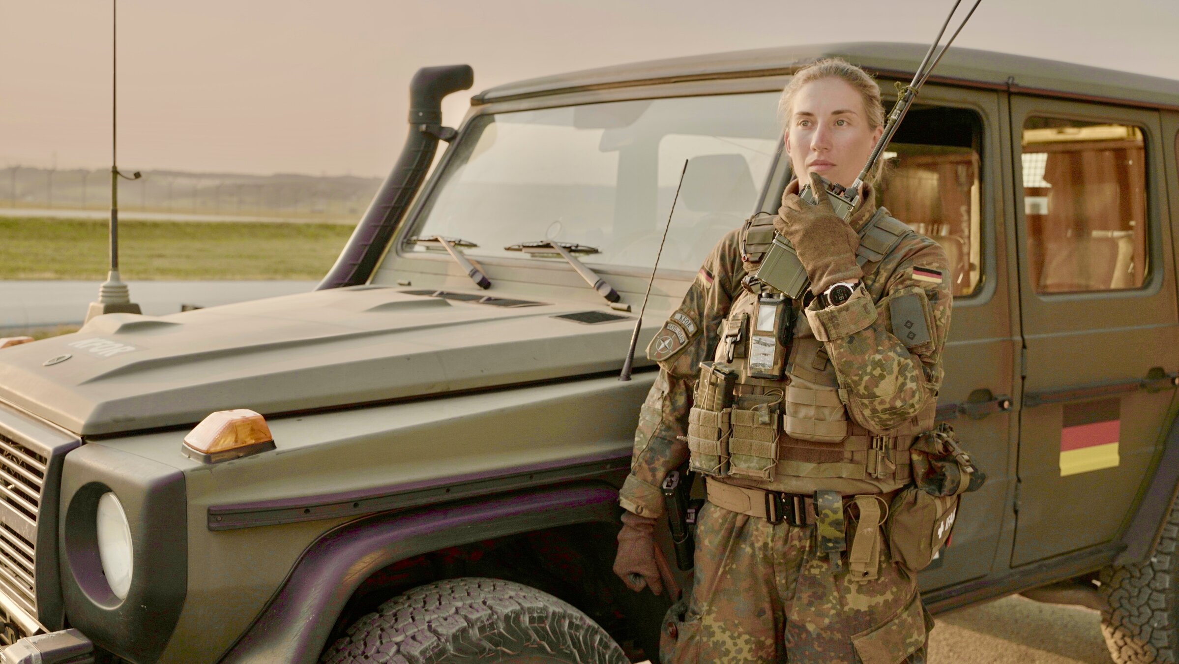 "37°: Hoher Einsatz: Soldaten im Kosovo": Alina steht in Flecktarn-Uniform und Schutzweste vor einem Bundeswehrauto. Sie hält das Handmikrofon eines Funkgeräts dicht an den Mund und spricht hinein.