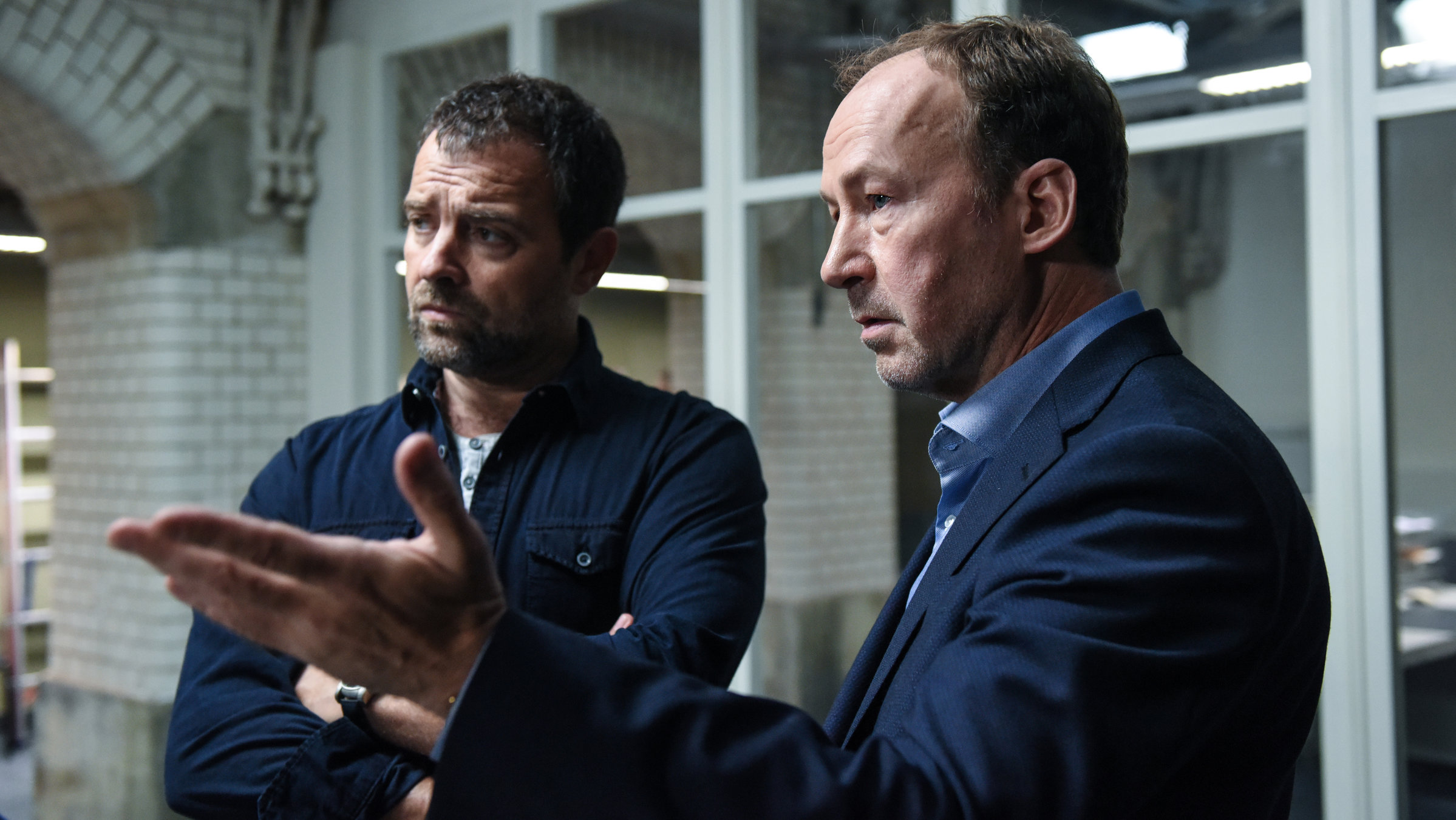 "Neben der Spur – Sag, es tut dir leid": Dr. Jessen (Ulrich Noethen) und Ruiz (Jürgen Maurer)stehen nebeneinander in einem Gebäude und schauen sich etwas an, auf das Dr. Jessen mit der linken Hand deutet. Ruiz hat die Arme vor der Brust verschränkt.
