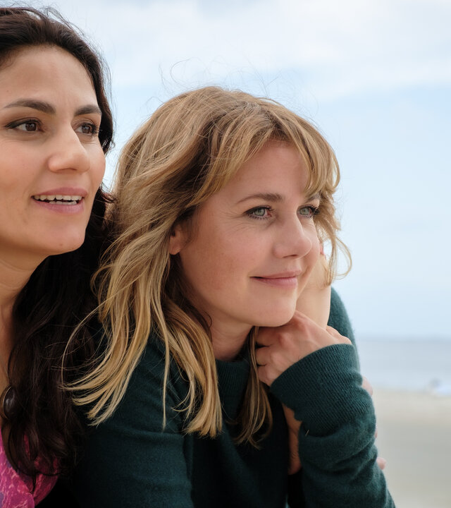 "Neuer Wind im Alten Land – Gestrandet": Beke (Felicitas Woll) und Elif (Halima Ilter) sitzen nebeneinander am Strand und blicken aufs Wasser.