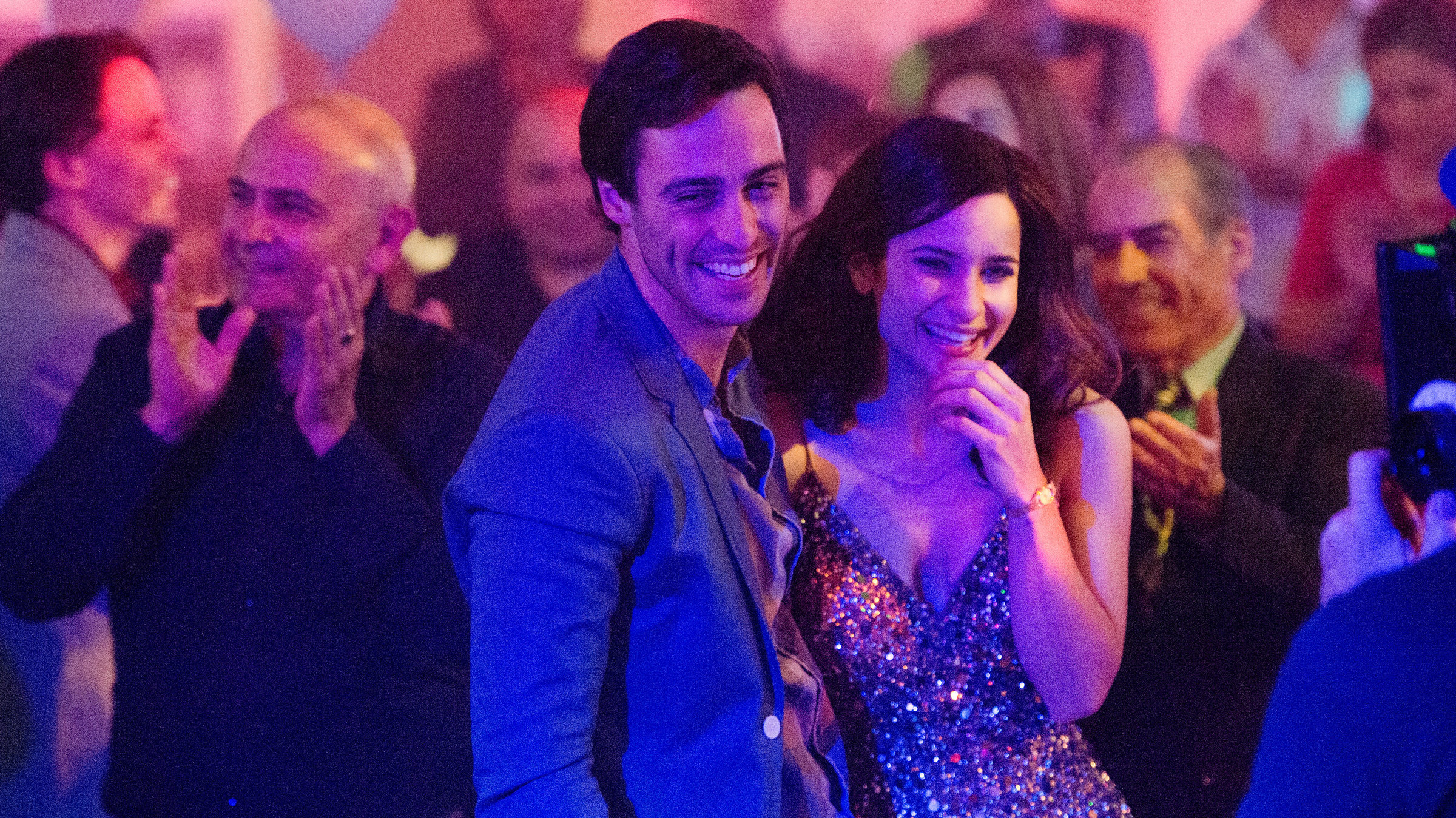 "Alex & Eve": Alex (Richard Brancatisano) und seine Freundin Eve (Andrea Demetriades) stehen lachend, Arm in Arm auf einem griechischen Fest. Im Hintergrund sind tanzende, fröhliche Menschen zu sehen.