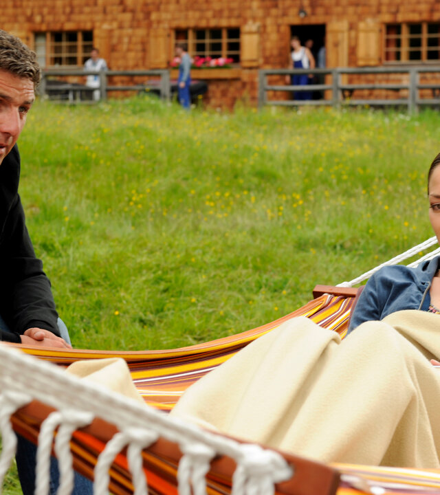 "Der Bergdoktor: Dunkle Wolken": Auf einer Wiese: Lisa Kleinert (Janina Flieger) liegt, in einer Decke gewickelt, in einer Hängematte. Sie hört aufmerksam Dr. Martin Gruber (Hans Sigl) zu, der neben ihr auf einem Stuhl sitzt.
