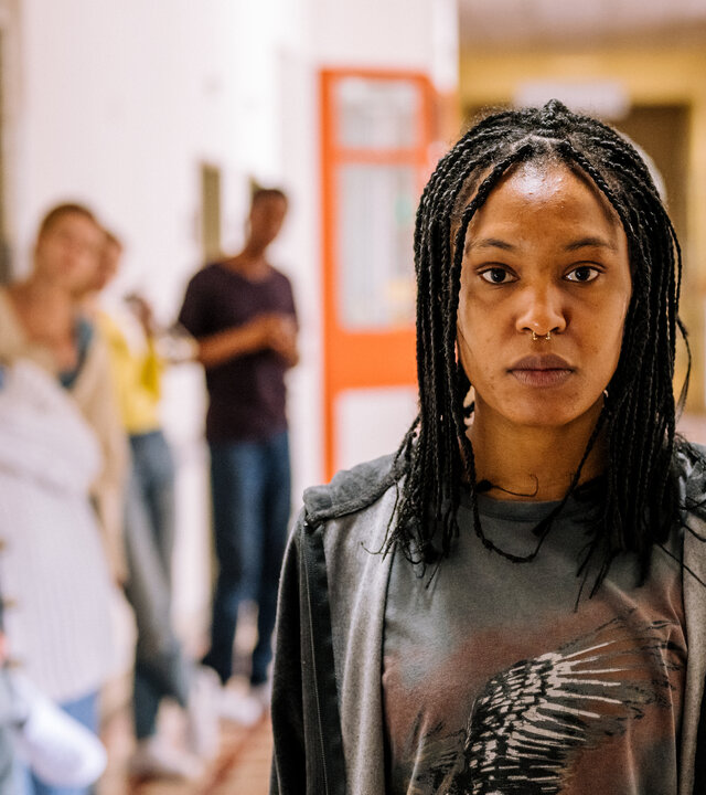 "Hungry - Trigger": Ronnie (Zoe Magdalena) steht auf dem Gang der Klinik und schaut emotionslos nach vorne. Hinter ihr lehnen, verschwommen zu erkennen, verschiedene Personen in einer Warteschlange an der Wand.