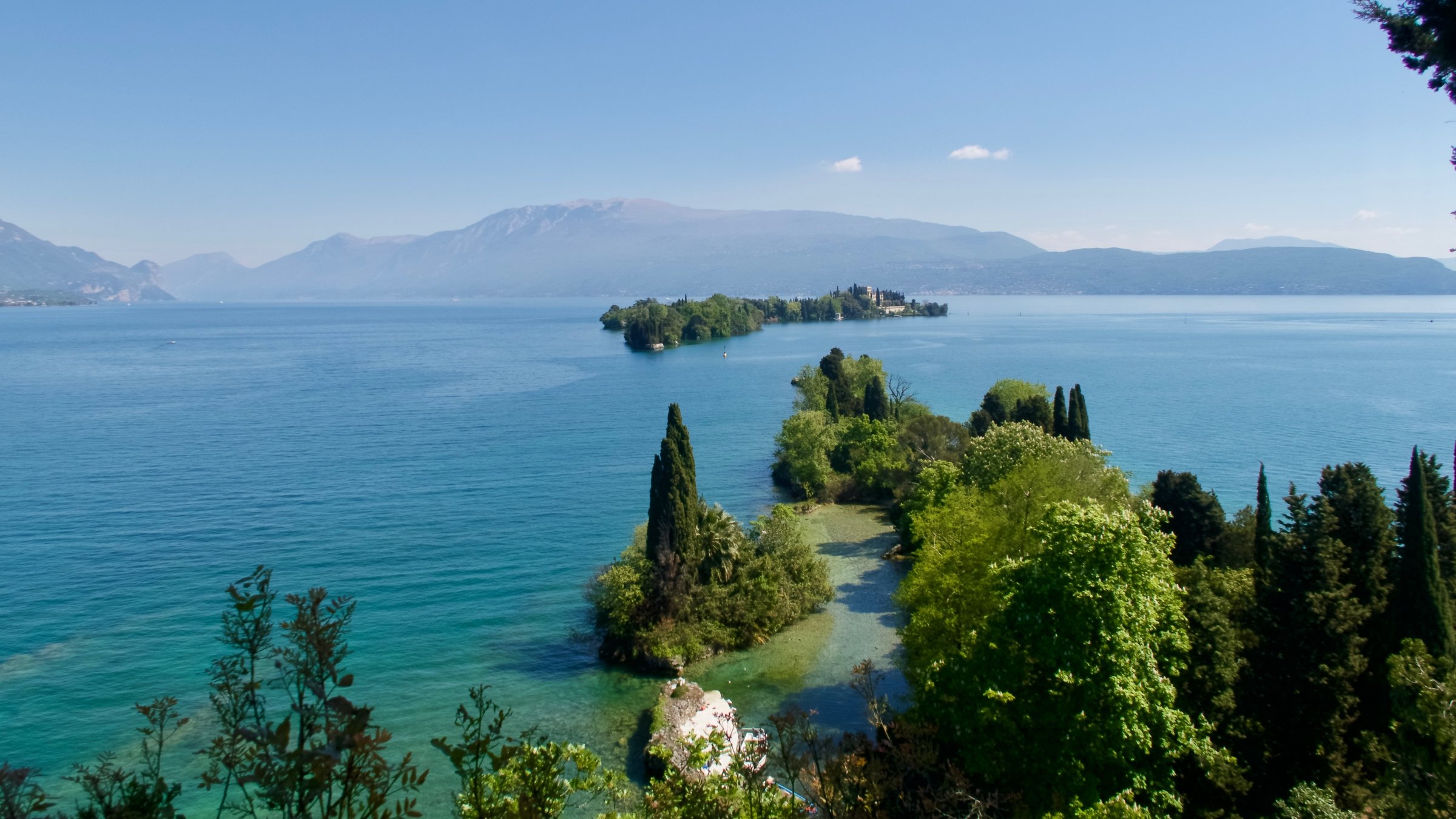 "Frühling am Gardasee - Sehnsucht nach dem Süden": Der Gardasee mit einer Insel in der Mitte, im Vordergrund Bäume.