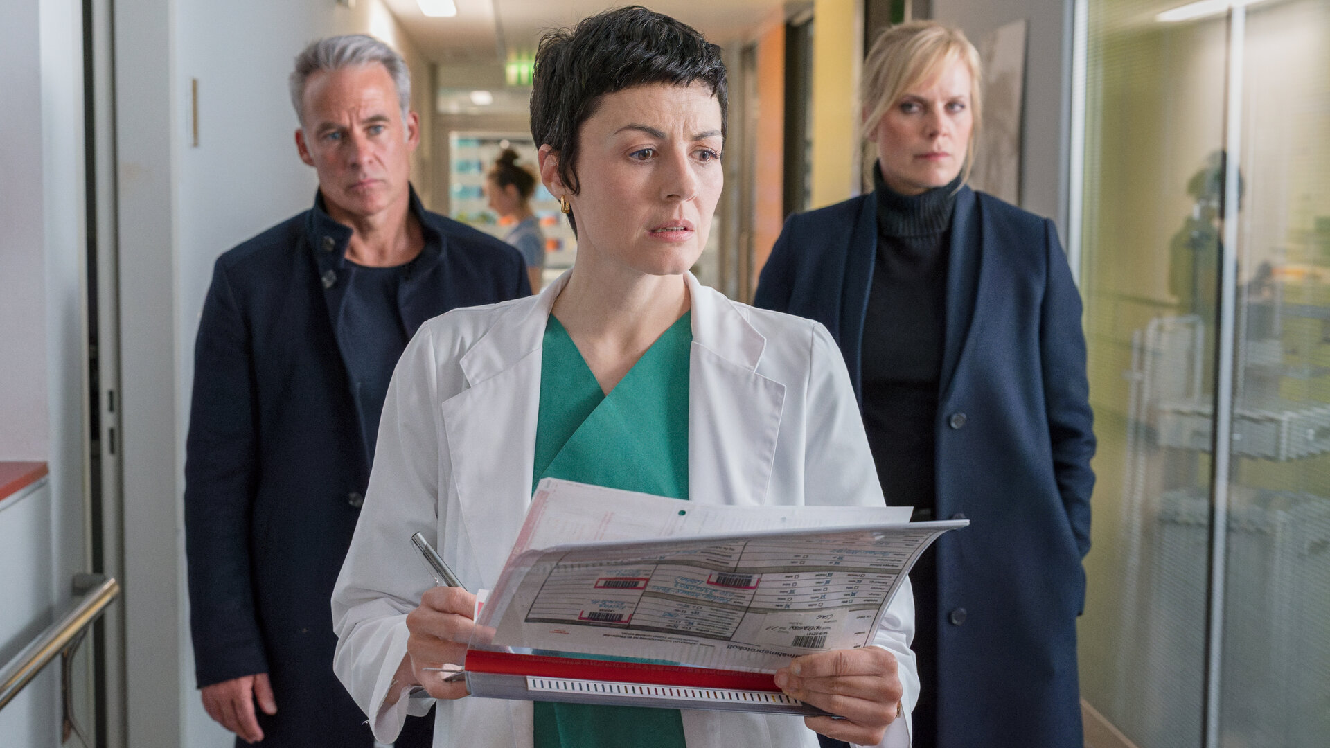 "SOKO Leipzig - In den Augen der Anderen": Jan Maybach (Marco Girnth) und Ina Zimmermann (Melanie Marschke) stehen hinter Antonia Kantor (Liza Tzschirner) in einem Krankenhausflur. Antonia Kantor trägt einen Arztkittel und hält Akten in den Händen.