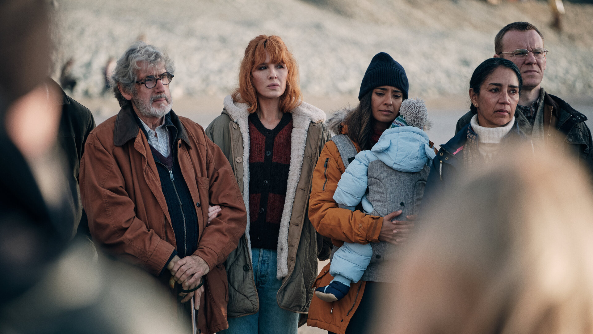 "Under Salt Marsh - Tod in den Salzwiesen - Rätselhafte Bilder": Owen (William Thomas), Jackie (Kelly Reilly), Nisha (Dinita Gohil), Ira (Seeta Indrani) und Mac (Brian Gleeson) stehen nebeneinander am Strand. Nisha trägt ein kleines Kind in einer Babytrage vor dem Körper.