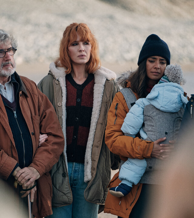 "Under Salt Marsh - Tod in den Salzwiesen - Rätselhafte Bilder": Owen (William Thomas), Jackie (Kelly Reilly), Nisha (Dinita Gohil), Ira (Seeta Indrani) und Mac (Brian Gleeson) stehen nebeneinander am Strand. Nisha trägt ein kleines Kind in einer Babytrage vor dem Körper.