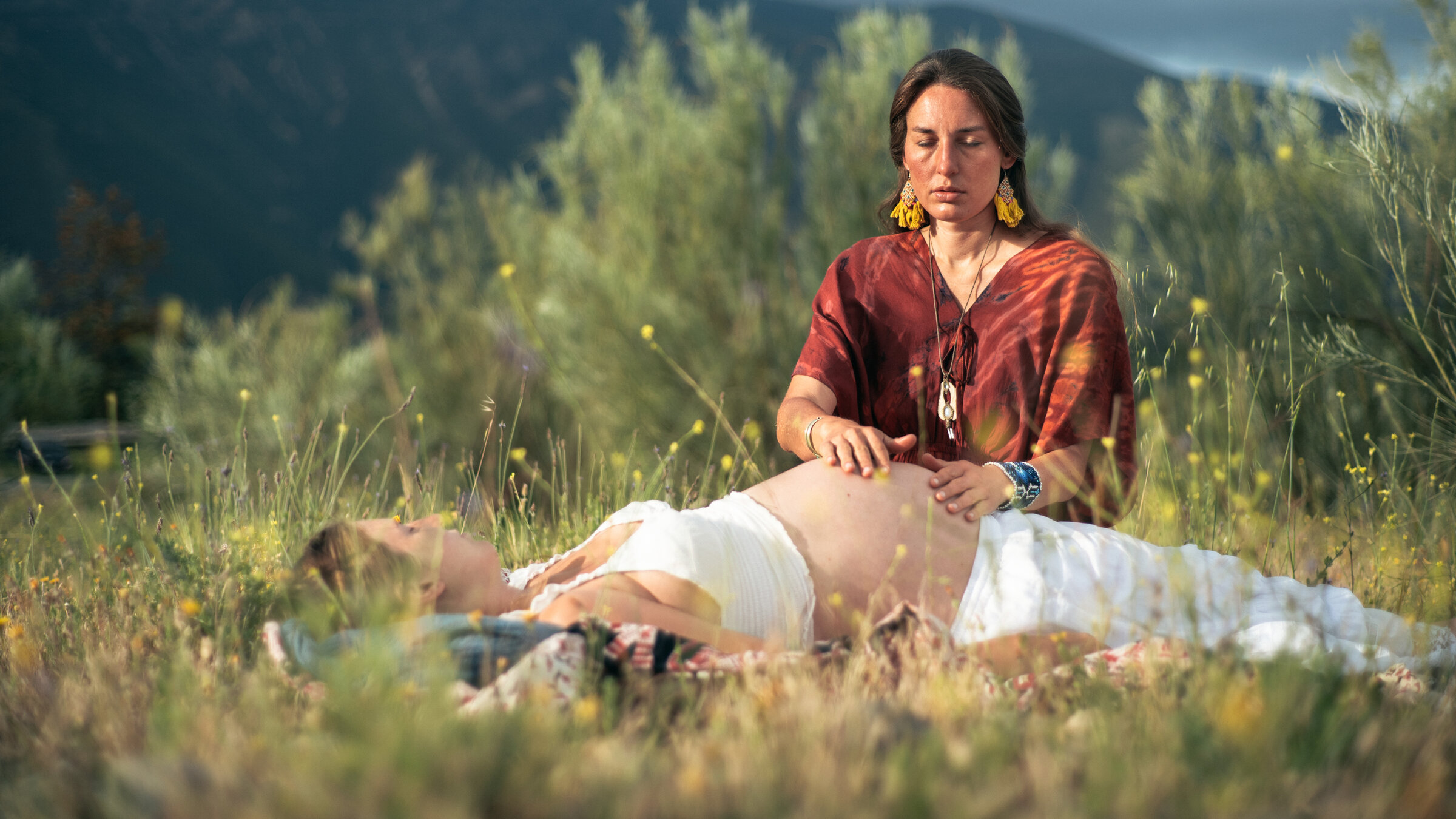 "37°Leben: Zwei Welten in mir": Eine Frau liegt im Gras, ihr Schwangerschaftsbauch ist nackt. Tiziana kniet daneben und gibt ihr eine Bauchmassage.