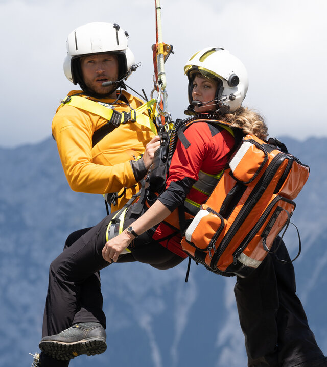 "Die Bergretter: Was wirklich zählt": Die Bergretter Katharina (Luise Bähr) und Markus (Sebastian Ströbel) hängen in ihrer Ausrüstung an einem Seil unter dem Rettungshubschrauber und schweben in der Luft. Im Hintergrund eine Berglandschaft.