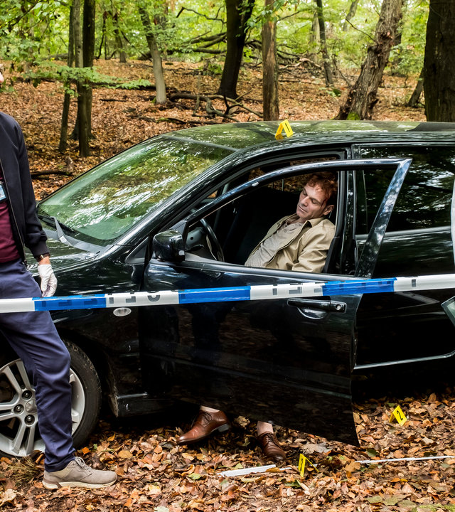 "SOKO Potsdam - Falsche Scham": Sophie Pohlmann (Katrin Jaehne) und Christoph Westerman (Hendrik von Bültzingslöwen) stehen am Tatort dem Spurensicherer Thomas Brandner (Yung Ngo) gegenüber. Thomas Brandner trägt einen weißen Overall, blaue Plastiküberzieher für die Schuhe und einen Mundschutz unter dem Kinn. Christoph hat einen Ausweis um den Hals hängen. Sophie Pohlmann trägt einen weißen Gummihandschuh an der rechten Hand. Neben den Kommissaren und dem Spurensicherer steht ein schwarzes Auto, auf dessen Fahrersitz der tote Andreas Littauer (Komparse) sitzt. Die Fahrertür ist geöffnet und seine Füße hängen aus dem Auto heraus. Der Tatort ist mit einem blau-weißen Absperrband gekennzeichnet.