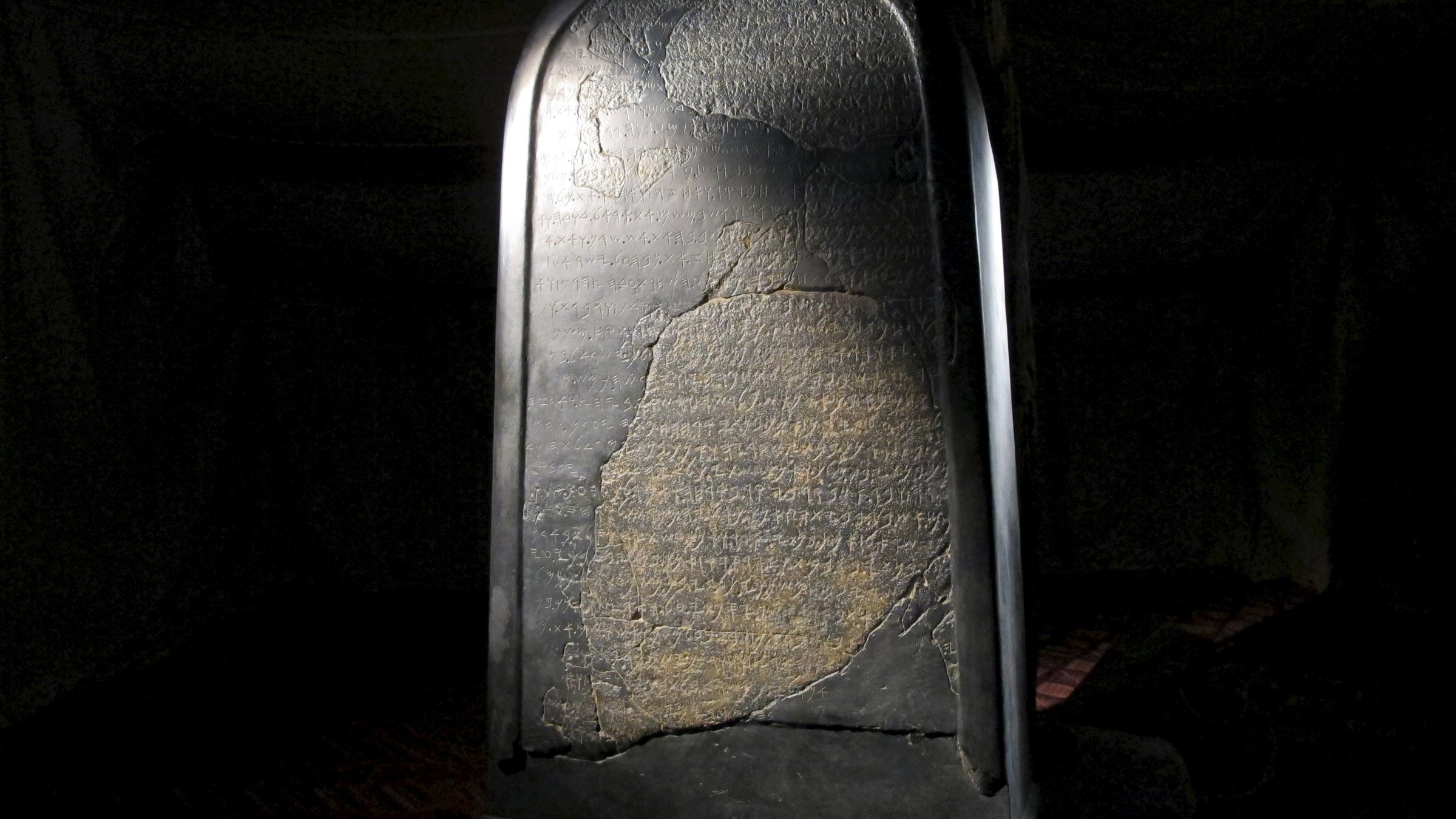 "Bibelrätsel - Die Karriere Gottes": Abguss der Mescha-Stele, 9. Jahrhundert vor Christus, auf der ein Text aus fremden Schriftzeichen zu sehen ist. Sie steht in einem dunklen Raum, angestrahlt von einem Licht.