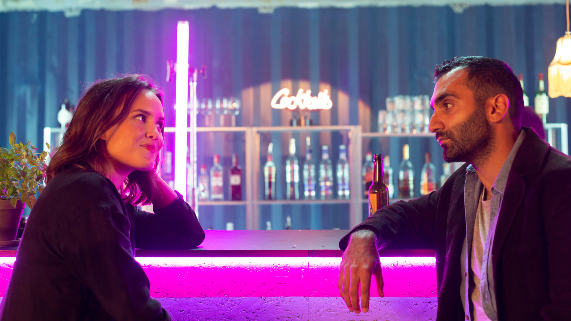 "Cryptid - Lisas Visionen": Lisa (Astrid Morberg) sitzt an einer Bar gegenüber von Lehrer Aron (Arvin Kananian). Beide lächeln sich an.